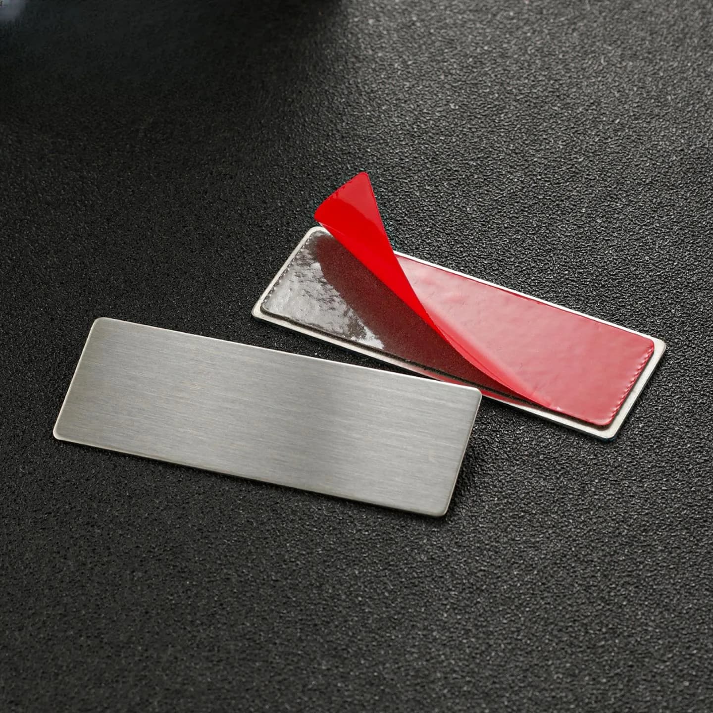 The back of a metal piece with a red 3M double-sided adhesive layer, the protective film partly peeled